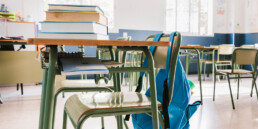 Classroom desk with books piled on top and a backpack hanging from the chair.